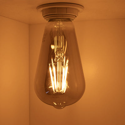 Close-up zijaanzicht van een brandende vintage LED filamentlamp in warm wit licht, gemonteerd in een plafondarmatuur, met zichtbare gloeidraden en amberkleurige gloed.