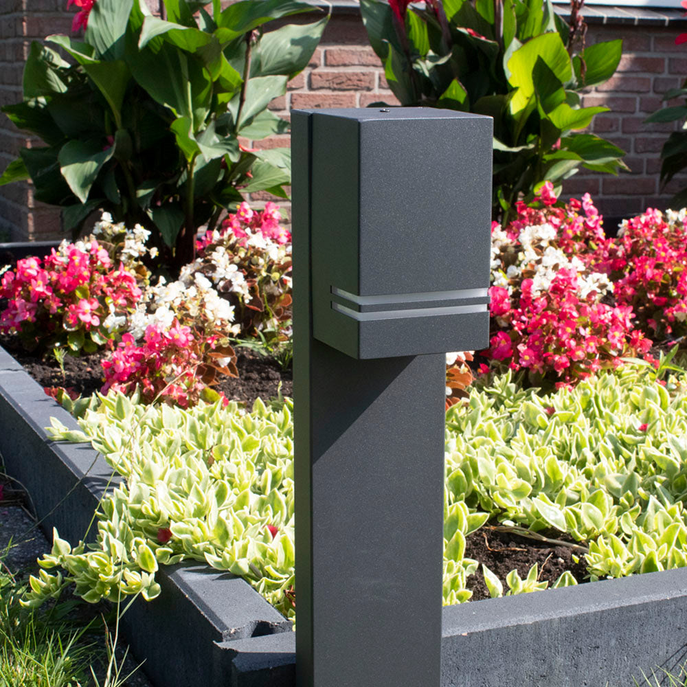 Moderne zwarte tuinlamp op sokkel, vooraanzicht, geplaatst langs een bloembed met kleurrijke bloemen en sierlijke bodembedekkers, ideaal voor sfeervolle tuinverlichting.