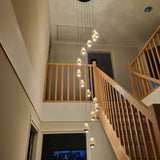 Moderne hanglamp met meerdere glazen lichtpunten in een verticale opstelling, gemonteerd in een hoge hal met houten trap en balustrade, gezien vanuit een laag perspectief.