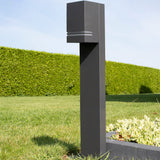 Moderne zwarte buitenlamp op paal met strakke vierkante vorm, gefotografeerd in zijaanzicht, geplaatst in een groene tuin met strakke haag op de achtergrond.