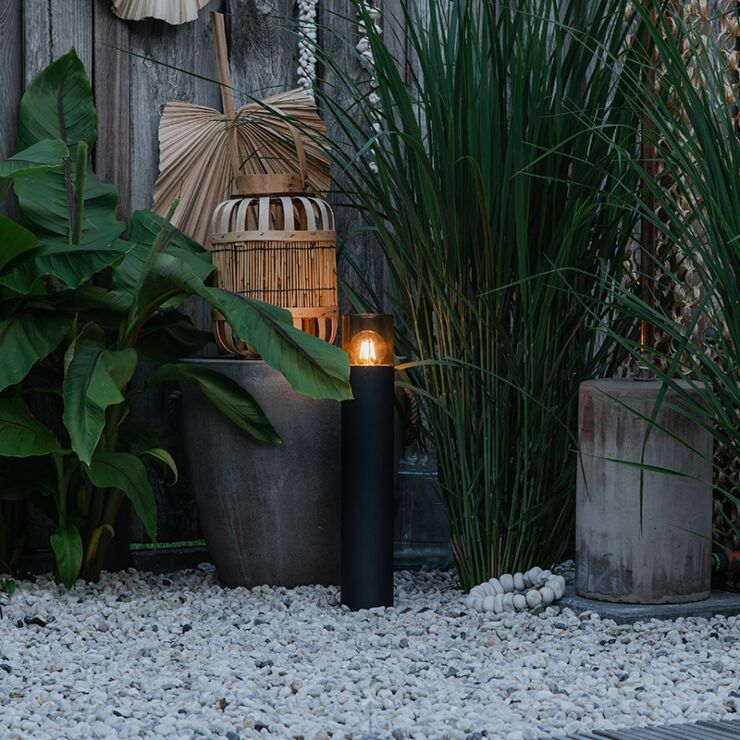 Zwarte moderne tuinlamp op zonne-energie in cilindervorm, zijaanzicht, geplaatst in een sfeervol ingerichte tuin met siergrind, grote plantenpotten en groene beplanting.