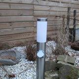 RVS buitenlamp met stopcontact in moderne tuin, frontaal aanzicht tegen houten schutting en siergrind.