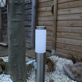 RVS buitenlamp met stopcontact, staande tuinlamp met cilindervormig design in zijaanzicht, geplaatst op witte sierstenen naast een boom en houten schutting.