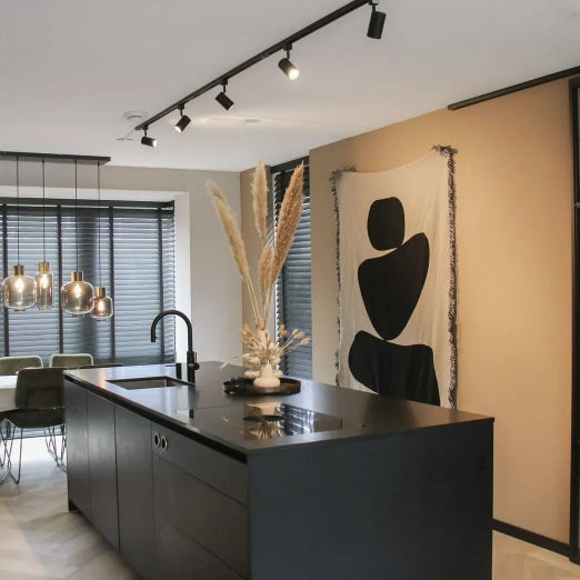 Moderne zwarte keukeneiland met inductiekookplaat en geïntegreerde spoelbak, gefotografeerd in een strak interieur met industriële hanglampen, houten jaloezieën en abstract wandkleed – frontaal zijaanzicht.