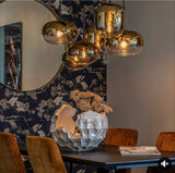 Close-up van een modern interieur met centraal een cluster van goudkleurige glazen hanglampen, gezien vanuit een zijaanzicht boven een eettafel met velvet stoelen, decoratieve vaas en donkerblauw behang met kraanvogels op de achtergrond.