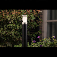 Moderne zwarte buitenlamp Rullo staand model, gefotografeerd in zijaanzicht in een tuinsetting met groene planten en paarse bloemen op de voorgrond.