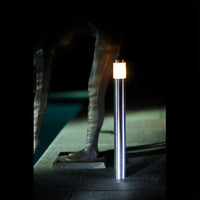 RVS buitenlamp met staand design en warme LED-verlichting, gefotografeerd in nachtelijke setting vanaf schuin vooraanzicht, geplaatst naast een kunstsculptuur bij een zwembad.