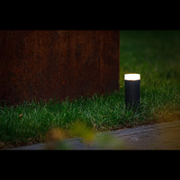 Moderne zwarte buitenlamp Philips Hue Calla sokkellamp, frontaal aanzicht in tuinsetting bij schemering, geplaatst langs een groen gazon en naast een roestkleurige muur.