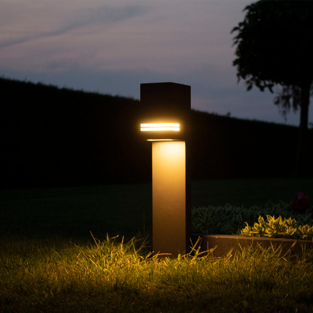 Moderne LED tuinlamp sokkelarmatuur in antraciet, sfeervol verlicht in een tuin tijdens de schemering, vooraanzicht.