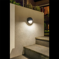 Ronde buitenwandlamp gemonteerd op een witte muur bij een buitentrap in de avond, met warm wit licht voor sfeervolle en functionele tuinverlichting – zijaanzicht.