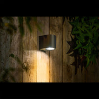 Moderne zwarte wandlamp voor buiten, gemonteerd op een houten schutting, gezien in zijaanzicht en verlichtend naar beneden voor sfeervolle tuinverlichting.