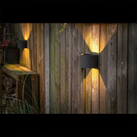 Zwart ronde buitenlamp met up- en downlight gemonteerd op een houten schutting, sfeervol verlichtend in een tuinsetting – zijaanzicht.